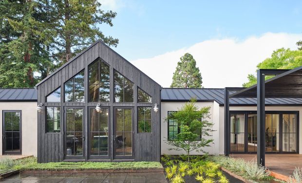 Exterior view of a contemporary single-storey residence with a prominent steep gable volume, dark vertical timber cladding, extensive multi-pane glazing, and adjoining low-pitched metal-roof sections. A covered outdoor deck and landscaped planting sit in front of the facade.