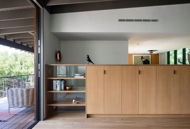 A contemporary open-plan living area with a low-profile built-in timber storage and display unit, featuring warm wood cabinetry, open shelving, books, and decorative objects. Large operable glazing opens onto a covered outdoor terrace with woven seating and leafy views. The interior has a restrained neutral palette, dark structural framing, timber flooring, and a partially visible dining or living zone beyond.