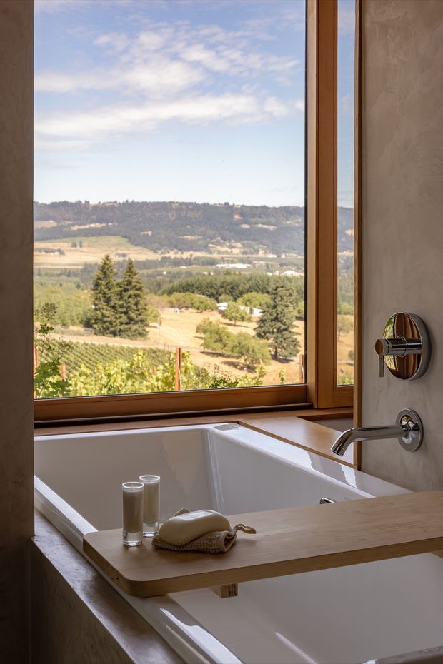 A serene bathroom with a freestanding white bathtub positioned beside expansive timber-framed windows. The windows overlook a sunlit rural vineyard and rolling wooded hills. A wall-mounted chrome bath filler and controls sit against a textured taupe plaster wall, while a timber bath tray holds glasses and a folded cloth. The space has a warm, natural, refined atmosphere with strong indoor-outdoor connection.