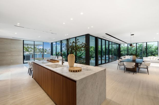 A spacious open-plan contemporary kitchen and living area with a large marble-look island, warm timber cabinetry, integrated seating, and extensive black-framed floor-to-ceiling glazing. The kitchen opens directly to a dining and lounge zone, with greenery visible outside and a bright, refined interior finish.