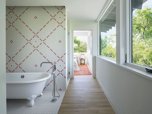Bright, elongated bathroom with a freestanding white clawfoot bathtub, wall-mounted bath filler, and a full-height geometric mosaic-tile feature wall in white with red patterned accents. Small-format pale floor tiles surround the tub, while the main circulation area has light timber flooring. Large windows run along the right side, bringing in abundant natural light and views of greenery. A doorway leads toward a furnished adjoining room.