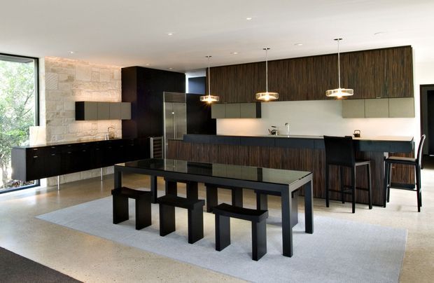 A spacious open-plan contemporary kitchen and dining area with dark timber cabinetry, black island and dining furniture, pale stone feature wall, and polished light flooring. Three pendant lights hang above the kitchen island, while a large dining table with bench seating occupies the foreground. Floor-to-ceiling glazing at left brings in abundant natural light and views of greenery.