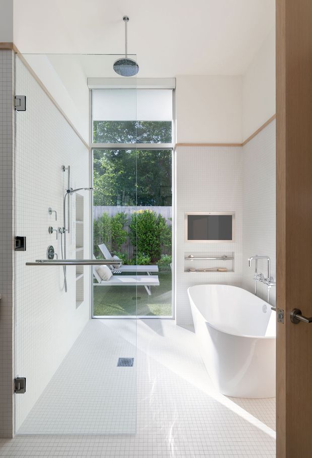 Bright contemporary bathroom with a frameless glass walk-in shower, white mosaic floor and wall tiles, freestanding white bathtub, wall-mounted television, open recessed shelving, and a large window overlooking a landscaped garden. Timber trim and a timber door add warmth to the predominantly white space.