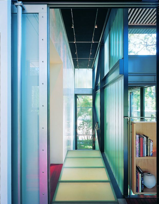 A narrow, light-filled interior passage or landing with translucent glass wall panels, exposed dark ceiling with track lighting, and strongly coloured blue-green structural and wall elements. A short set of broad steps leads toward floor-to-ceiling glazing and greenery outside. Open shelving with books and objects is visible to the right, while a red upholstered or painted surface appears at the left.