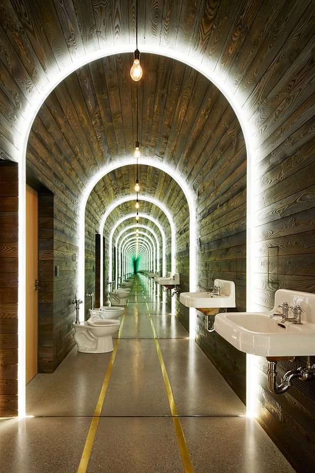 A dramatic, highly repetitive bathroom corridor with a long perspective view. The space features arched illuminated openings, dark timber wall and ceiling cladding, polished concrete or terrazzo-like flooring with parallel yellow floor lines, exposed plumbing, and repeated white wall-mounted sinks and toilets. Bare pendant bulbs hang along the vaulted passage, creating a theatrical, industrial atmosphere.