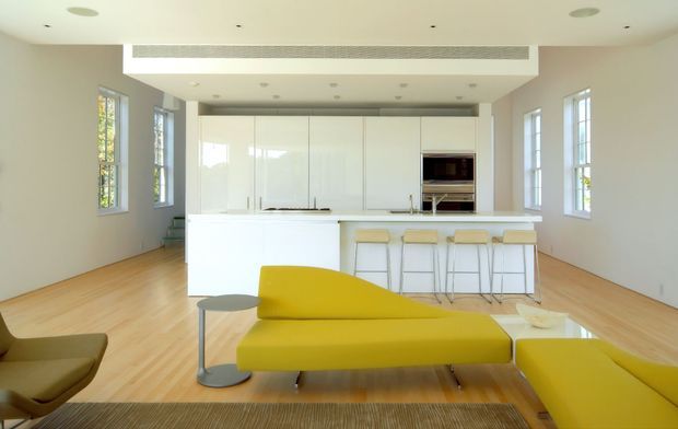 Bright open-plan contemporary interior with a minimalist white kitchen, large island with integrated seating, pale timber flooring, tall multi-pane windows, and a sculptural mustard-yellow sofa and ottoman in the foreground. The space has a clean, airy layout with a raised ceiling section and recessed lighting.