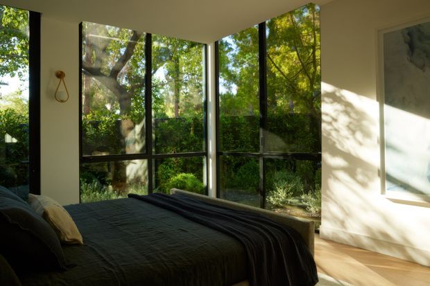 A serene bedroom with a low-profile bed dressed in dark charcoal bedding, set against floor-to-ceiling black-framed glazing overlooking lush greenery. Warm sunlight creates strong dappled shadows across the pale walls and timber floor. A minimal wall light and partially visible artwork complete the understated, nature-connected interior.