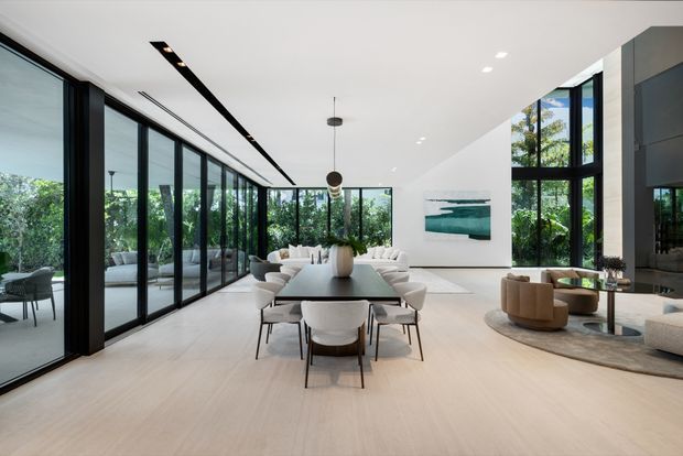 A spacious, high-end open-plan interior combining a dining area and living room. A long dark dining table with upholstered chairs sits centrally beneath suspended pendant lights, while a pale sectional sofa and curved lounge chairs form a seating area beyond. Full-height black-framed glazing lines both sides of the room, opening views to lush greenery. The space has pale timber flooring, white walls and ceiling, dark built-in wall elements, contemporary artwork, and abundant natural light.