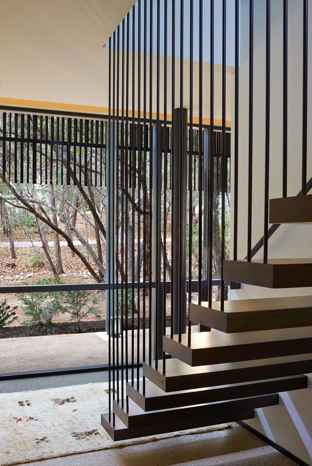 Interior architectural photograph focused on a sculptural floating staircase. Dark timber or stained-wood treads are suspended within a tall open stairwell by slender black vertical metal balusters, with a minimalist dark handrail. Full-height glazing opens views to a dry, wooded landscape outside. The space has crisp white walls and ceiling, a pale neutral floor, and strong linear geometry with abundant natural light.