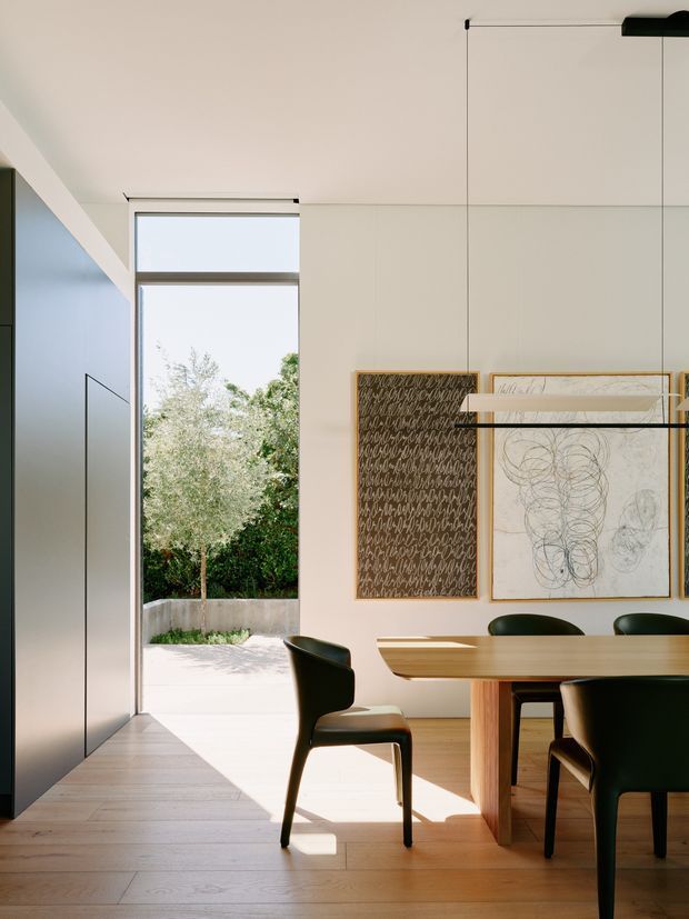 A bright, refined dining area in a contemporary home, viewed toward a tall open doorway leading to a small courtyard. The room has pale timber flooring, white walls and ceiling, a large sculptural timber dining table, dark green upholstered dining chairs, minimalist black-framed glazing and a linear pendant light. Abstract artwork is displayed on the wall, while integrated dark joinery is visible at the left.