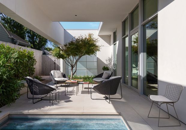 A contemporary courtyard terrace beside a small swimming pool, furnished with four dark woven lounge chairs and a light wire-frame chair around low outdoor tables. The space is enclosed by a white rendered building with extensive dark-framed glazing, a high covered soffit, and a rectangular skylight/opening. Pale large-format paving, a planted tree, and boundary screening create a private, minimalist outdoor entertaining area.