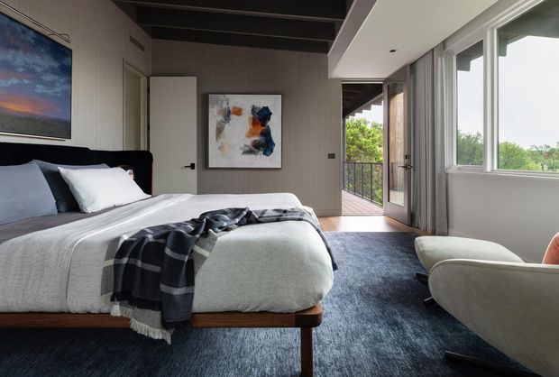 A contemporary bedroom with a neatly made bed, dark timber bed frame, layered white and grey bedding, and a dark patterned throw. The room features textured grey wall covering, abstract artwork, a dark exposed ceiling with beams, and large windows opening toward a balcony with leafy views. A pale upholstered lounge chair sits in the foreground on a deep blue-grey carpet.