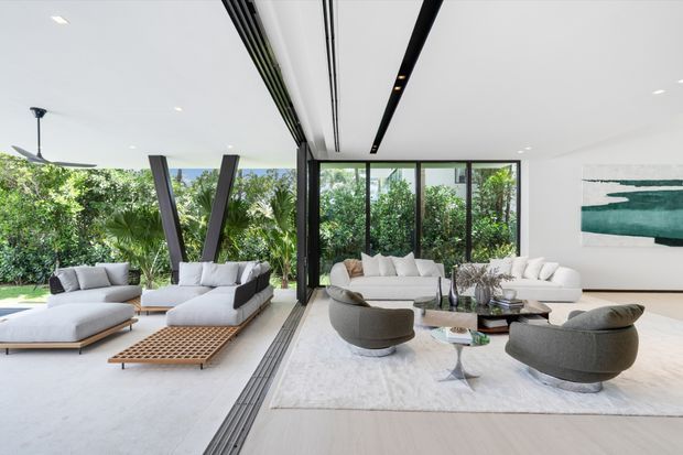 Bright, spacious contemporary open-plan living room with extensive floor-to-ceiling glazing opening toward lush greenery. The room features pale flooring, neutral upholstered sofas and sculptural lounge chairs, a large light rug, minimalist recessed and linear ceiling lighting, dark structural or window framing, and abstract teal artwork. A ceiling fan and outdoor foliage are visible at the edges.