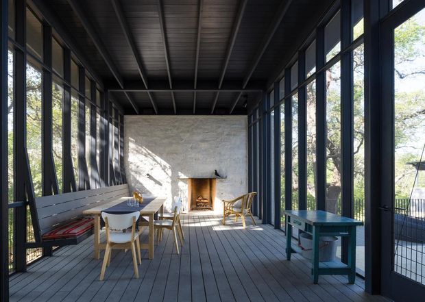 A long, enclosed sunroom or screened outdoor living space with full-height dark-framed glazing on both sides, a timber plank floor, exposed dark ceiling rafters, built-in bench seating, a dining table and chairs, and a masonry fireplace at the far end. The room opens visually to a wooded setting and is filled with natural light, creating a calm indoor-outdoor connection.