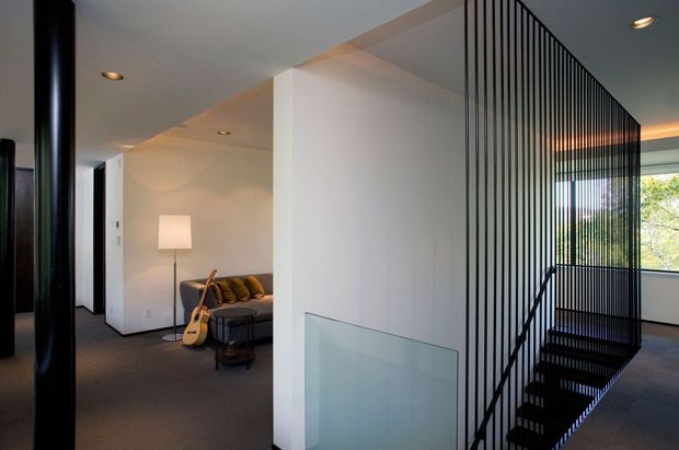 Interior view of a contemporary home featuring a dark carpeted floor, white walls and ceiling, recessed lighting, and a dramatic black metal open-riser staircase enclosed by a vertical black slat screen. A small sitting area with a sofa, floor lamp, and guitar is visible beyond the central wall, while a large window brings in views of greenery.