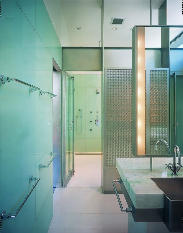 A compact, highly contemporary bathroom with pale turquoise glass wall panels, integrated chrome towel rails and hooks, a glass-enclosed shower, and a large mirror. A broad pale stone or engineered-stone vanity with an undermount basin and sculptural chrome tap occupies the foreground. A perforated or textured translucent privacy screen separates the vanity from the shower area, while recessed ceiling fixtures and a linear, minimalist composition create a clean modern aesthetic.