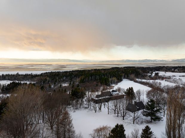 Aerial view of a snow-covered rural or coastal property surrounded by leafless deciduous trees and evergreen woodland. Several dark-roofed buildings are scattered across the landscape, with expansive tidal flats or water and distant hills visible beyond. The scene is captured under dramatic winter clouds with warm light near the horizon.