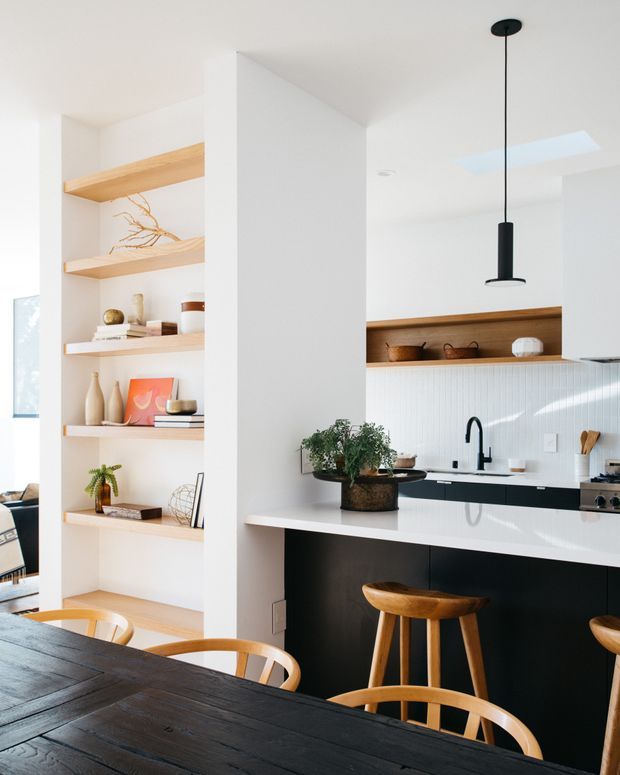 Bright open-plan kitchen and dining area with a minimalist white interior, black island cabinetry and pendant light, white vertically tiled splashback, timber open shelving, and a dark timber dining table with curved wooden chairs. A skylight and large window provide abundant natural light, while plants, ceramics, and decorative objects add warmth.