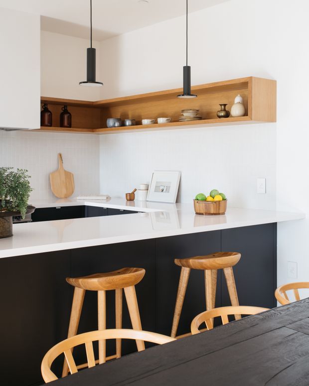 Bright contemporary kitchen with a white waterfall-edge island, matte black base cabinetry, and a warm timber open shelving unit wrapping the upper corner. Two black pendant lights hang over the island, while timber stools and a dark dining table with light timber chairs occupy the foreground. White tiled backsplash, simple ceramics, and natural timber accessories create a clean, warm, understated interior.