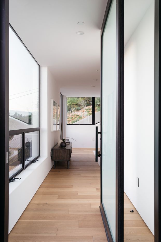 A bright, narrow modern hallway with full-height glazing and black-framed internal glass doors. The corridor has pale timber flooring, white walls and ceiling, a dark timber console, and views toward a planted sloping landscape through the windows. The space is clean-lined and minimally furnished, with strong natural light and a reflective glass partition on the right.