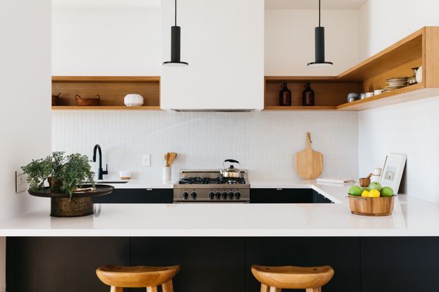 Bright contemporary kitchen with a large white island and integrated seating, matte black lower cabinetry, white vertical-tile splashback, warm timber open shelving, and white upper cabinetry. Two black pendant lights hang above the island, while the rear wall includes a black tap, cooktop and range, open shelving, and kitchen accessories.