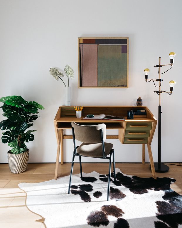 A stylish home office vignette with a mid-century-inspired wooden writing desk, open shelving, drawers, and a modern upholstered chair. The desk is arranged against a light wall beneath an abstract artwork, accompanied by a sculptural multi-arm floor lamp, potted tropical plant, books, stationery, and a black-and-white cowhide rug over timber flooring. The space is bright, refined, and warm with a vintage-modern character.