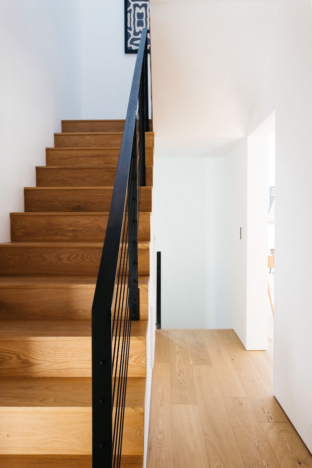Bright contemporary interior view of a timber staircase with broad oak-toned treads and matching light timber flooring. A slender black metal balustrade with vertical cable infill runs up the stair, contrasting with the white walls and clean-lined hallway. Natural light enters from the right, and a small patterned decorative panel is visible near the stair landing.