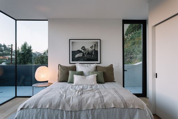 A contemporary bedroom with a neatly made bed positioned against a pale wall, layered in white and muted olive-green cushions and a softly striped grey throw. Full-height black-framed glazing wraps the left corner, while a black-framed glazed door opens to an exterior balcony or terrace. A framed black-and-white animal photograph hangs above the bed, and a warm spherical bedside lamp sits on a small round table. The room has light timber flooring, minimalist built-in storage, and a calm, neutral atmosphere.