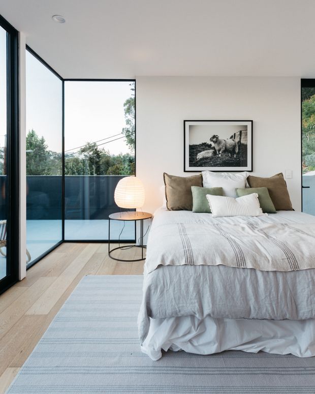 Bright contemporary bedroom with a neatly styled bed, layered neutral and pale green bedding, a small round black side table with a softly glowing lamp, pale timber flooring, and a large striped rug. Floor-to-ceiling black-framed glazing opens to a balcony or exterior view, bringing in abundant natural light. A framed black-and-white sheep photograph hangs above the bed.