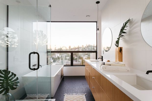 Bright contemporary bathroom with a freestanding bathtub beside a large window, a glass-enclosed shower, and a long timber-veneer vanity with white countertop and integrated basins. White tiled walls, dark small-format floor tiles, rounded mirrors, black tapware, and indoor tropical plants create a clean, refined interior with city views.