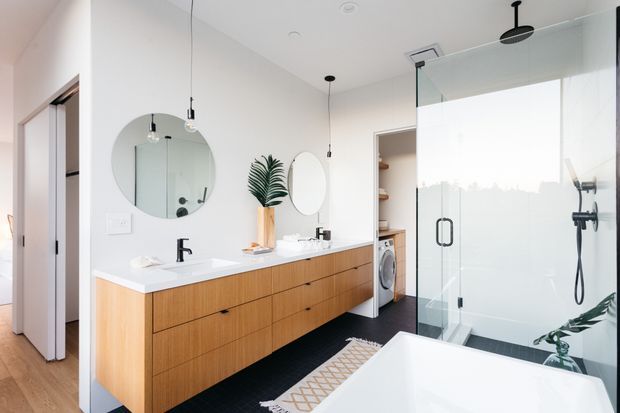 Bright contemporary bathroom with a long floating timber vanity featuring two white basins and black tapware, round mirrors, pendant lights, and a large frameless glass shower enclosure. A freestanding white bathtub occupies the foreground, with a laundry appliance visible in an adjacent utility area. The room uses white walls and surfaces, warm timber joinery, dark flooring, and minimal greenery.