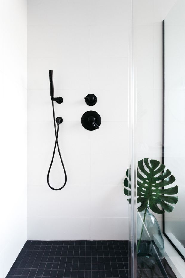 Minimalist walk-in shower with large white wall tiles, matte black shower fittings and a dark tiled floor. A clear glass partition encloses the shower, with a large monstera leaf in a glass vase visible beyond it. The space is bright, clean and high-contrast.