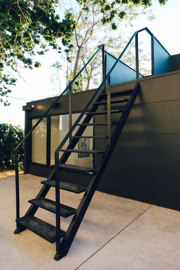 Exterior view of a compact modern building with a steep black metal open-riser stair leading to an elevated rooftop or upper-level terrace. The dark metal stair and railings contrast with smooth dark wall cladding, translucent or glazed upper panels, and a pale concrete paved ground. Mature trees surround the structure, creating a leafy outdoor setting.