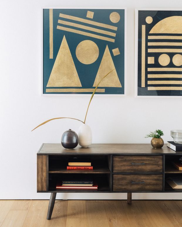 A styled contemporary living room vignette featuring a low dark timber sideboard with open shelving and drawers, displayed against a white wall. Geometric blue-and-gold artwork hangs above the console, which is decorated with ceramic vases, a small plant, books, and a glass vessel. Pale timber flooring and tapered legs create a warm, mid-century-inspired look.