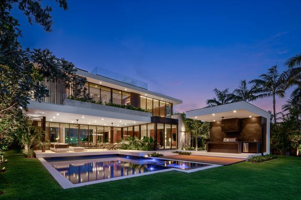 A high-end contemporary two-storey residence photographed at dusk, with extensive floor-to-ceiling glazing, projecting flat rooflines, vertical screening, landscaped gardens, and a large reflective swimming pool. A separate covered outdoor entertaining pavilion includes a built-in kitchen or bar area and opens onto a timber-toned deck.