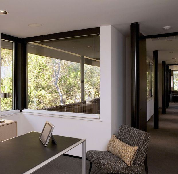 A modern, light-filled interior with a compact desk or worktable positioned beside expansive windows overlooking leafy vegetation. A textured upholstered lounge chair with a cushion sits in the foreground, while a long corridor with repeated black cylindrical posts and glazed openings extends into the background. White walls, dark trim, recessed lighting, and neutral finishes create a calm contemporary workspace.