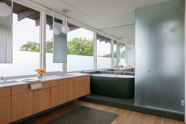 A bright, modern bathroom with a long timber vanity and multiple basins along the left wall, large windows, a freestanding dark bathtub in the corner, and a translucent glass privacy partition on the right. The room features white surfaces, dark small-format floor tiling around the bath platform, timber flooring in the foreground, globe pendant lights, and views of greenery through the windows.