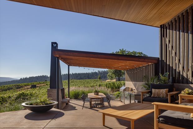 A contemporary covered outdoor terrace overlooking landscaped grounds and vineyard-like planting. The space features a timber-lined roof, dark vertical slatted cladding, a timber-framed pergola with integrated lighting, concrete paving, outdoor lounge furniture, a low timber coffee table, dining seating, and a large planter. The open setting provides expansive views toward forested hills under a clear sky.
