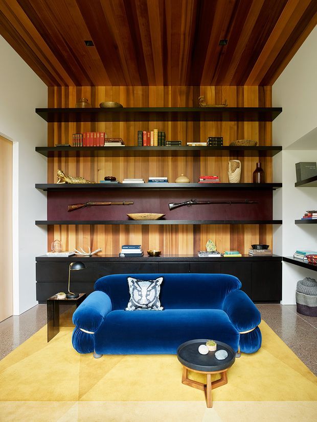A richly detailed living room centered on a vivid blue velvet sofa and round coffee table, set against a full-height timber-lined feature wall with dark floating display shelves, curated books and decorative objects, and a long black built-in cabinet. The ceiling continues the warm timber panelling, with recessed downlights. A large golden-yellow area rug and terrazzo-like floor add contrast, while displayed firearms and sculptural objects give the room a curated, eclectic character.