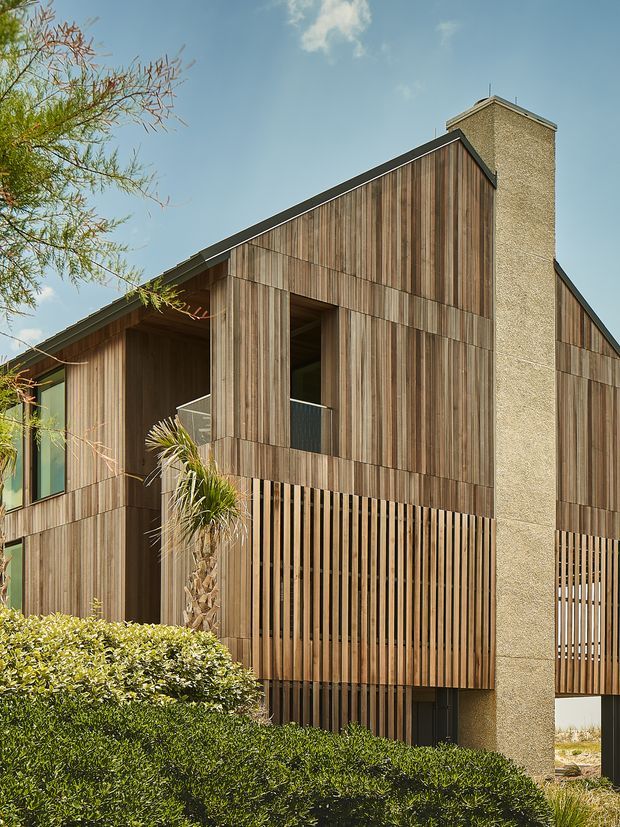 Exterior view of a contemporary multi-storey building with steep gabled roof forms, extensive vertical timber cladding and screened openings, plus a tall textured masonry chimney or structural wall. Landscaped hedges and palms sit in the foreground under a clear sky.