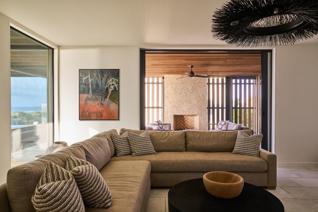 A warm, contemporary living room with a large taupe L-shaped sectional sofa, textured cushions, and a dark rounded coffee table. The room opens through wide sliding glazing to a covered outdoor area with a timber-lined ceiling, textured fireplace wall, and vertical screening. Coastal views are visible through the side glazing. A dramatic black sculptural pendant light hangs overhead, while pale walls and large light floor tiles create a bright, relaxed setting.