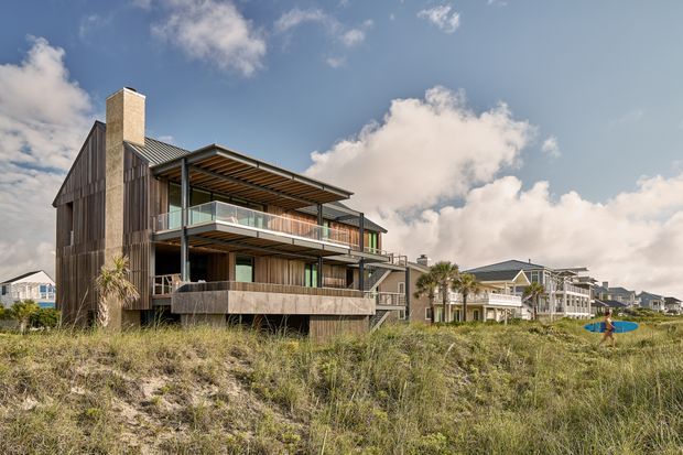 Exterior architectural photograph of a contemporary multi-level coastal residence set among sandy dunes. The home features dark vertical cladding, extensive glazing, broad sheltered balconies, timber-toned soffits, a projecting upper-level terrace, and a tall masonry chimney. Similar coastal residences extend along the shoreline in the background.