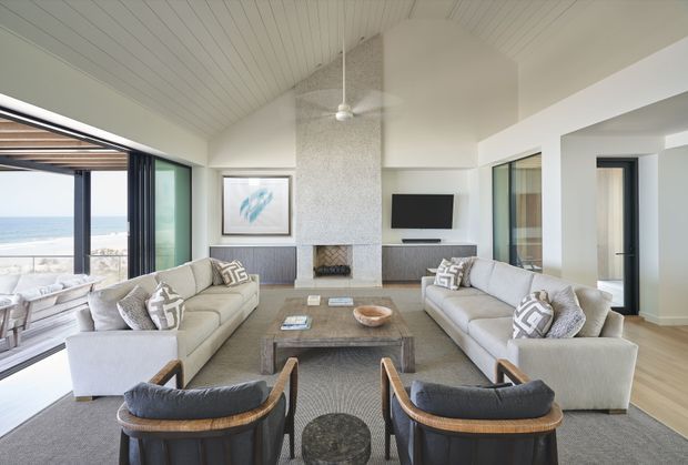 Bright coastal living room with a large sectional sofa arrangement facing a textured stone fireplace and wall-mounted television. The room has a white painted plank ceiling with a ceiling fan, light timber flooring, neutral upholstered seating, dark wood-toned built-in cabinetry, and extensive glazing opening toward an ocean view. Two barrel-back lounge chairs and a low central coffee table complete the seating area.