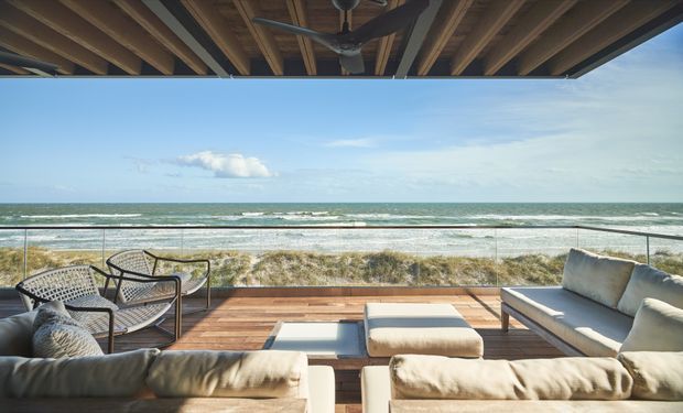 A covered outdoor lounge terrace overlooking a sandy beach and ocean. The space features a timber deck, exposed timber ceiling structure with a ceiling fan, glass balustrade, and a large arrangement of neutral upholstered sofas, ottomans, and woven lounge chairs.