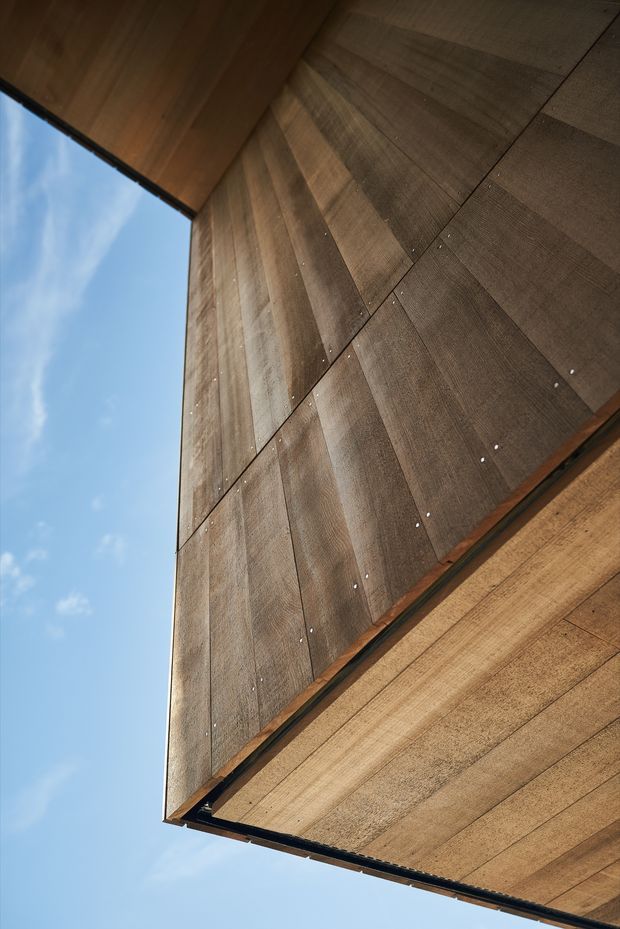 Upward-looking close-up of a modern building facade clad in warm brown wood-toned panels, with visible panel joints and small metal fixings. A projecting roof or soffit frames the facade against a clear blue sky, emphasizing the angular contemporary construction.