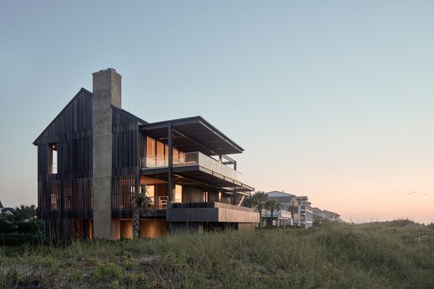 Exterior architectural photograph of a contemporary multi-level coastal house set among dune vegetation. The building features dark vertical timber-like cladding, a tall exposed masonry chimney, extensive glazing, projecting balconies, and broad flat roof overhangs. Similar homes recede along the shoreline in the background under soft evening light.