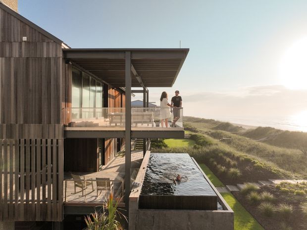 Exterior architectural photograph of a multi-level coastal home with dark timber cladding, broad projecting roof planes, extensive glazing, and layered balconies overlooking dune vegetation and the ocean. A narrow reflecting or swimming pool runs alongside the building, with an elevated terrace and outdoor seating visible.