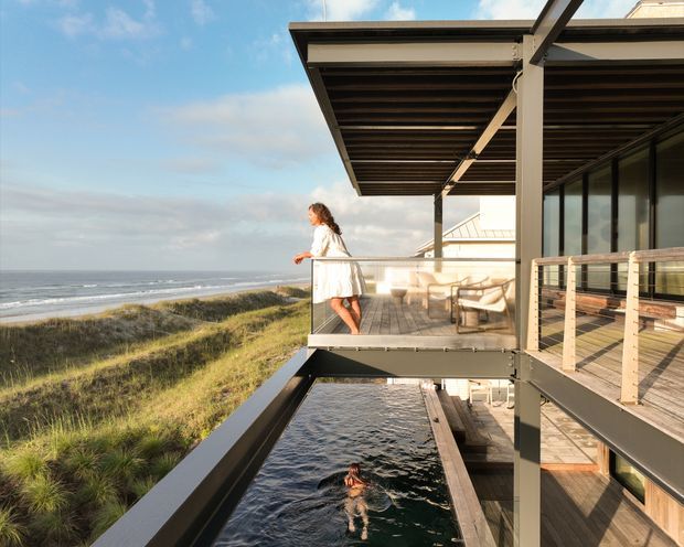 A modern coastal house on an elevated structure overlooking a beach and ocean. The image shows a timber-decked balcony with glass and timber railings, a covered outdoor seating area, broad roof overhangs, and a narrow swimming pool below the deck. Large glazed openings connect the exterior balcony to the building interior. A person stands on the balcony and another swims in the pool.