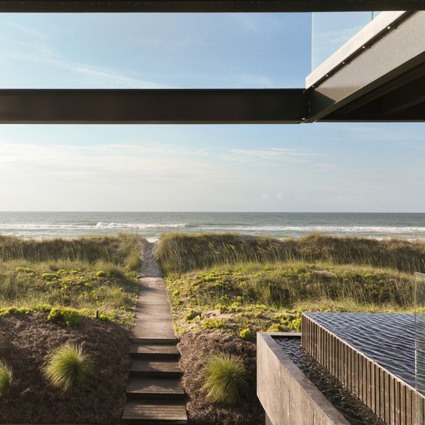 View toward the ocean from a modern coastal property, with a narrow timber boardwalk winding through sandy dunes to the beach. Dark horizontal roof or pergola beams frame the upper view, while a built-in pool or water feature with vertical timber screening occupies the lower right. The scene combines contemporary architecture with natural dune planting and expansive coastal views.