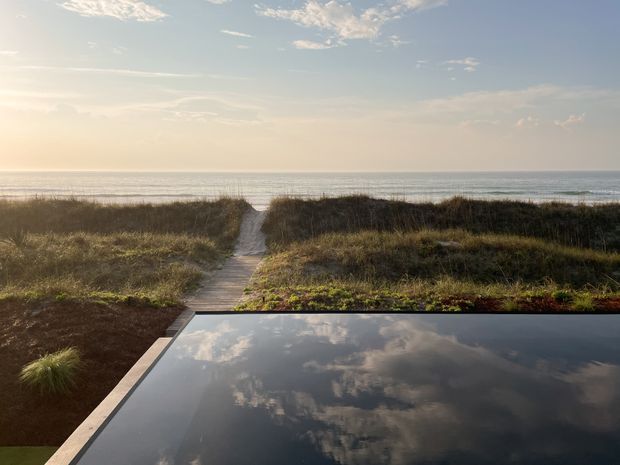 A tranquil coastal landscape viewed from a property, with a narrow timber boardwalk leading through low dune vegetation to the beach and ocean. In the foreground is a dark reflective water surface, likely a pool, mirroring the partly cloudy sky. Warm low-angle sunlight creates a serene, natural atmosphere.